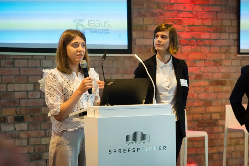 15 Jahre IEGUS: Nora Schulte-Coerne und Katharina Kirstein während ihrer Input-Präsentation. Katharina Kirstein hält ein Mikrofon in der Hand und spricht. Nora Schulte-Coerne blickt zu ihr herüber.