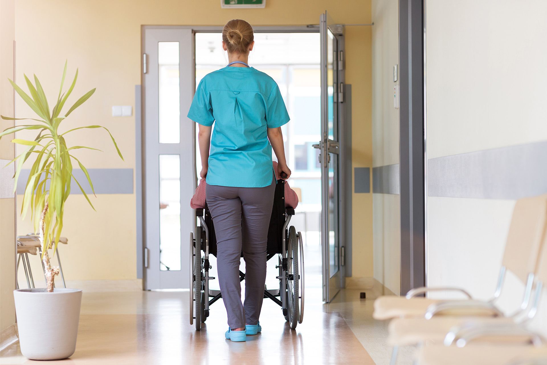 Ausbildungsabbrueche in der Pflege verhindern: Krankenschwester in blaugrünem Kasack schiebt eine Frau im Rollstuhl durch einen Krankenhausflur.