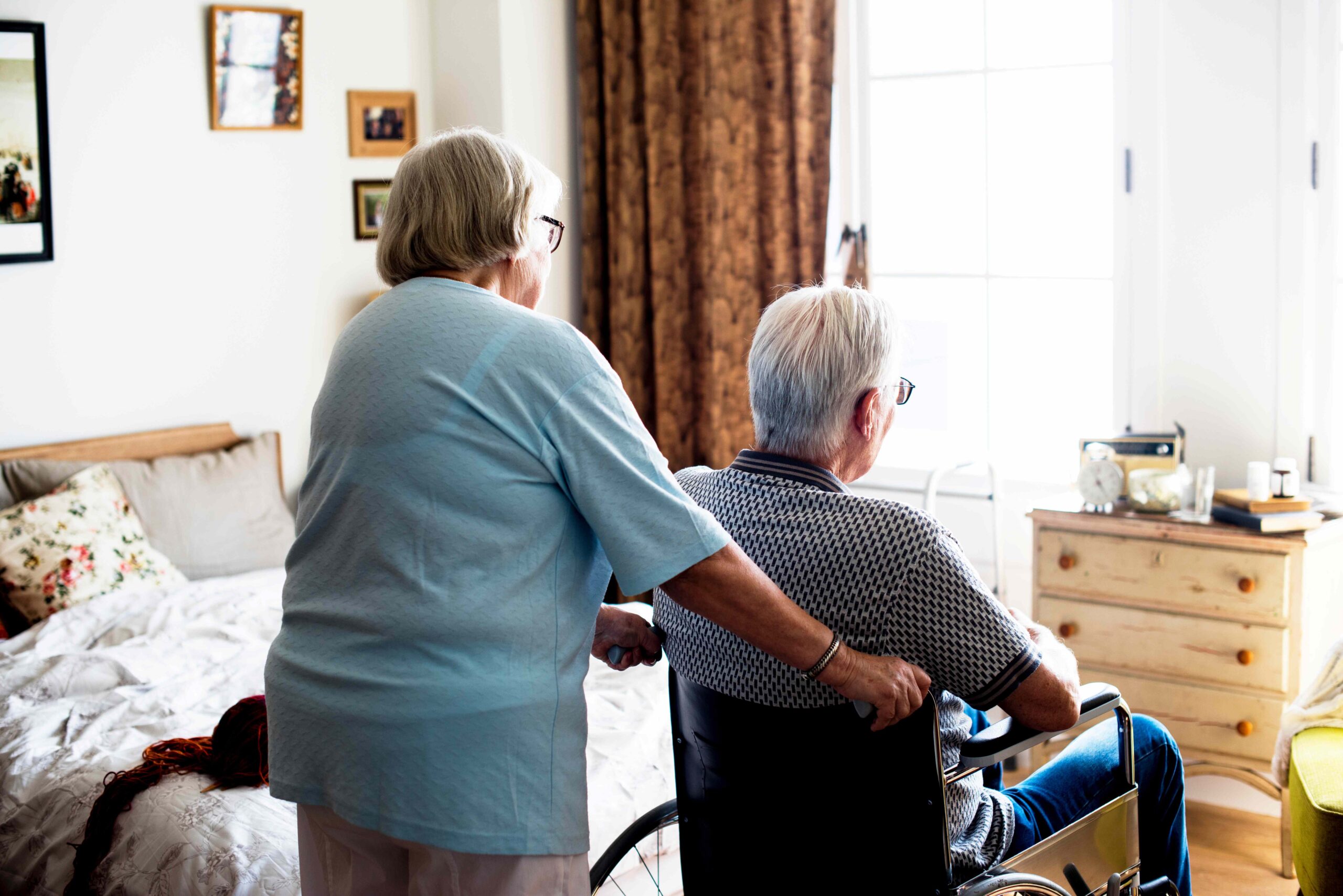 Die unsichtbare Last: Gesundheitliche Auswirkungen informeller Pflege: Das Bild zeigt ein älteres Paar in einem Schlafzimmer. Eine in blau gekleidete Frau schiebt einen im Rollstuhl sitzenden Mann durch das Zimmer.